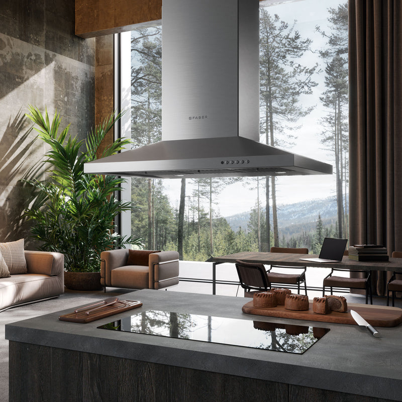Modern kitchen and living area with large windows overlooking a forest. The kitchen features a stainless steel Faber range hood above an island with an integrated cooktop.