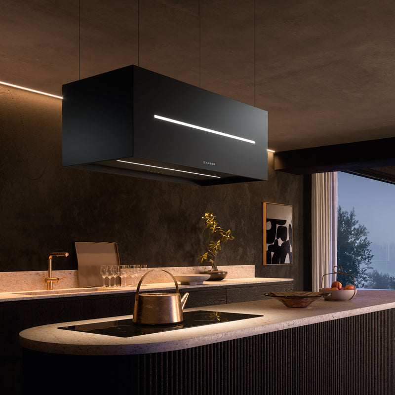 Modern kitchen featuring a sleek, black Graziosa range hood suspended above a light-colored island countertop. The island includes a built-in stovetop, a copper kettle, a bowl of fruit, and glassware. The kitchen has dark walls, ambient lighting, and a window with partially drawn curtains