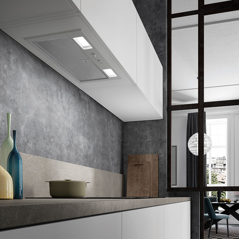 faber inca smart gray lifestyle image of a painted light gray insert kitchen range hood with 2 square led lights and one long mesh filter and slider controls. The kitchen features a textured gray backsplash, a sleek countertop with decorative bottles, and a cooking pot. In the background, a dining area with contemporary decor and a large window is visible.