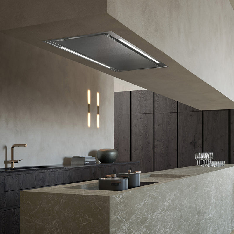 Industrial grey kitchen featuring a long marble island with an integrated stove top and Stratus Isola range hood in ceiling over it.